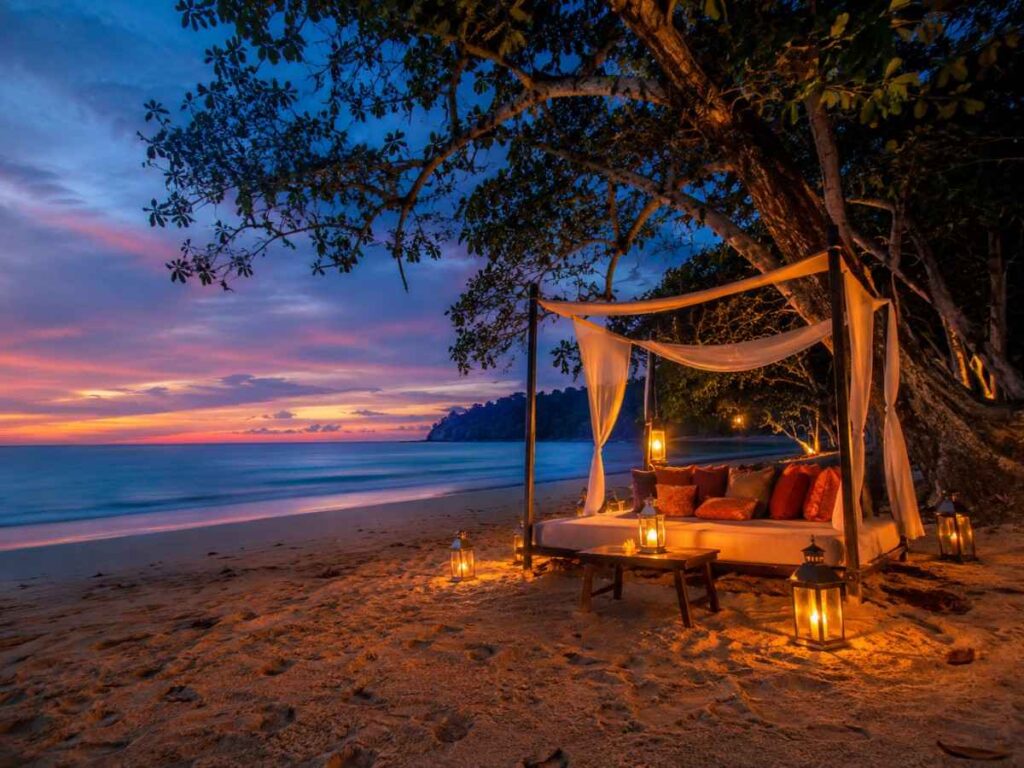 A secluded luxury lounge setup with lanterns and white linen on the sand at Radhanagar Beach, Andaman Islands, during twilight after a quiet sunset.