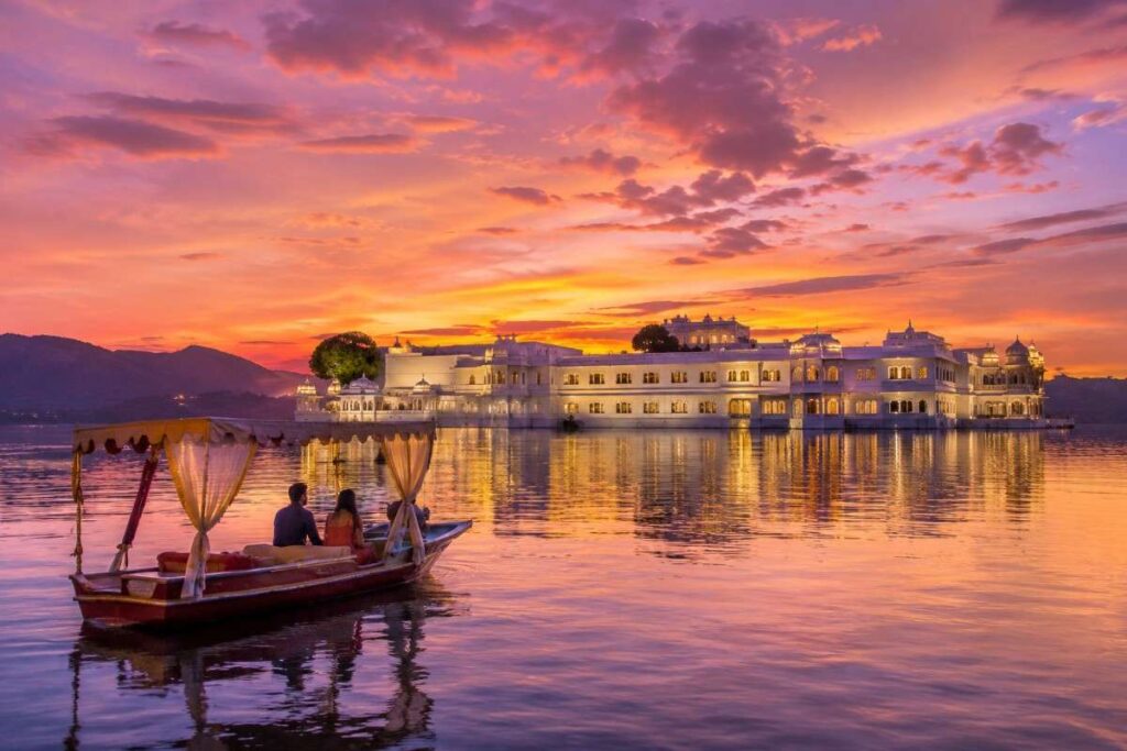 A wide view of the Taj Lake Palace in Udaipur reflecting in Lake Pichola during a gold and pink sunset, with a small traditional shikara boat in the water.